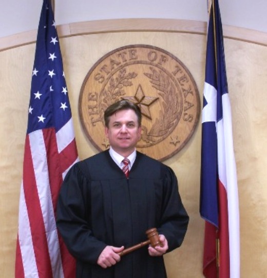 Wharton County Judge Phillip Spenrath standing in front of the State of Texas Seal, the United State Flag and the Texas State Flag
